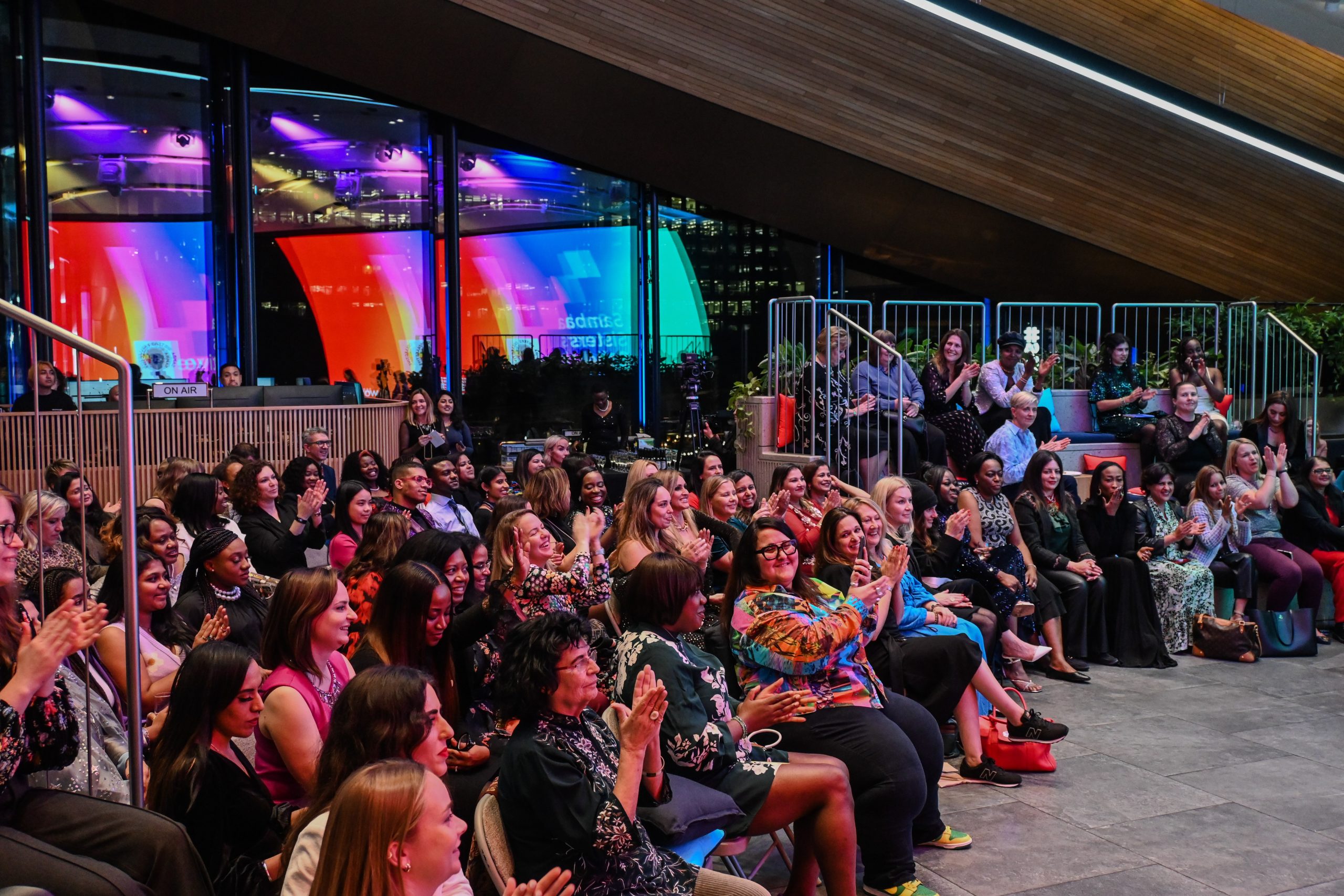 An audience, lit by multi-coloured lights, sit watching the UN Women UK Awards Ceremony with delight on their faces.