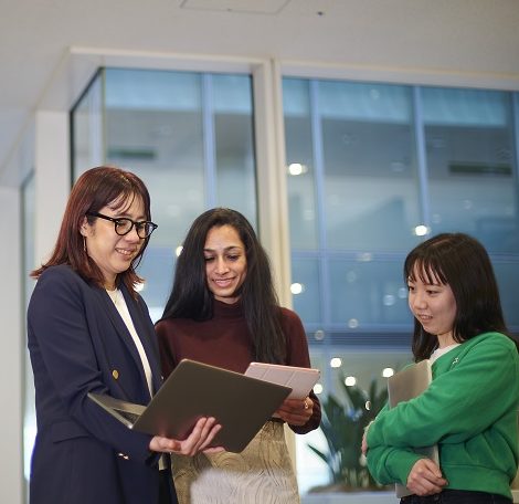 Three women in an office environment, review work on a laptop.