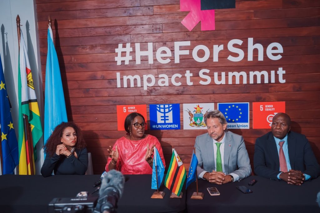 Three women and a man sit at a trestle table in front of a wooden backdrop that reads '#HeForShe Impact Summit'